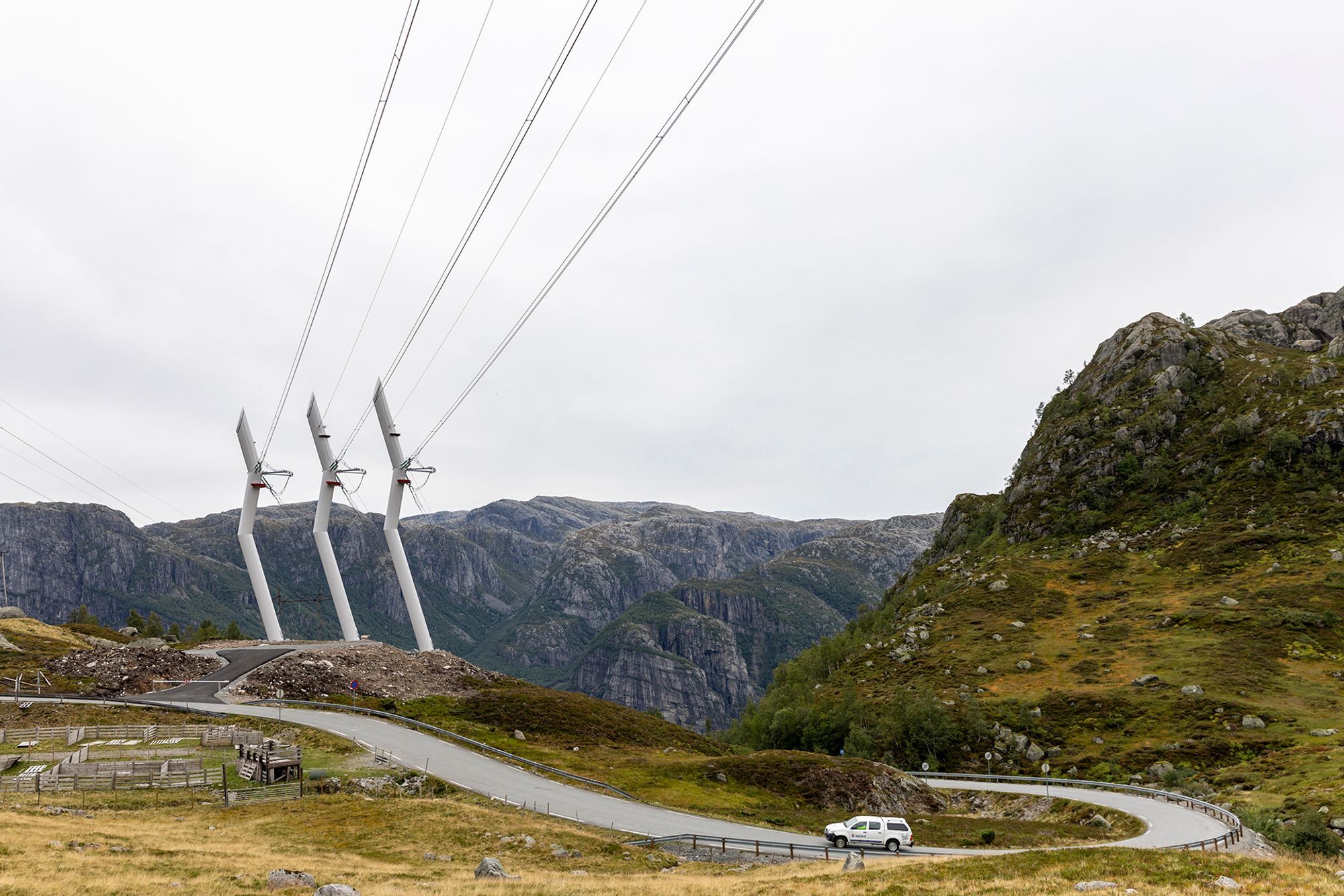 Three single transmission towers in the middle of hills and a white car cruising on a road next to it