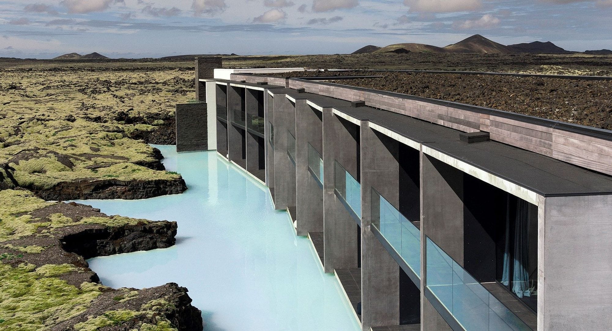 The concrete structure of the Blue Lagoon Hotel seen in the distance with the light blue water of the lagoon along the building in a flurry of moss-covered lava fields and mountains and blue sky in the distance