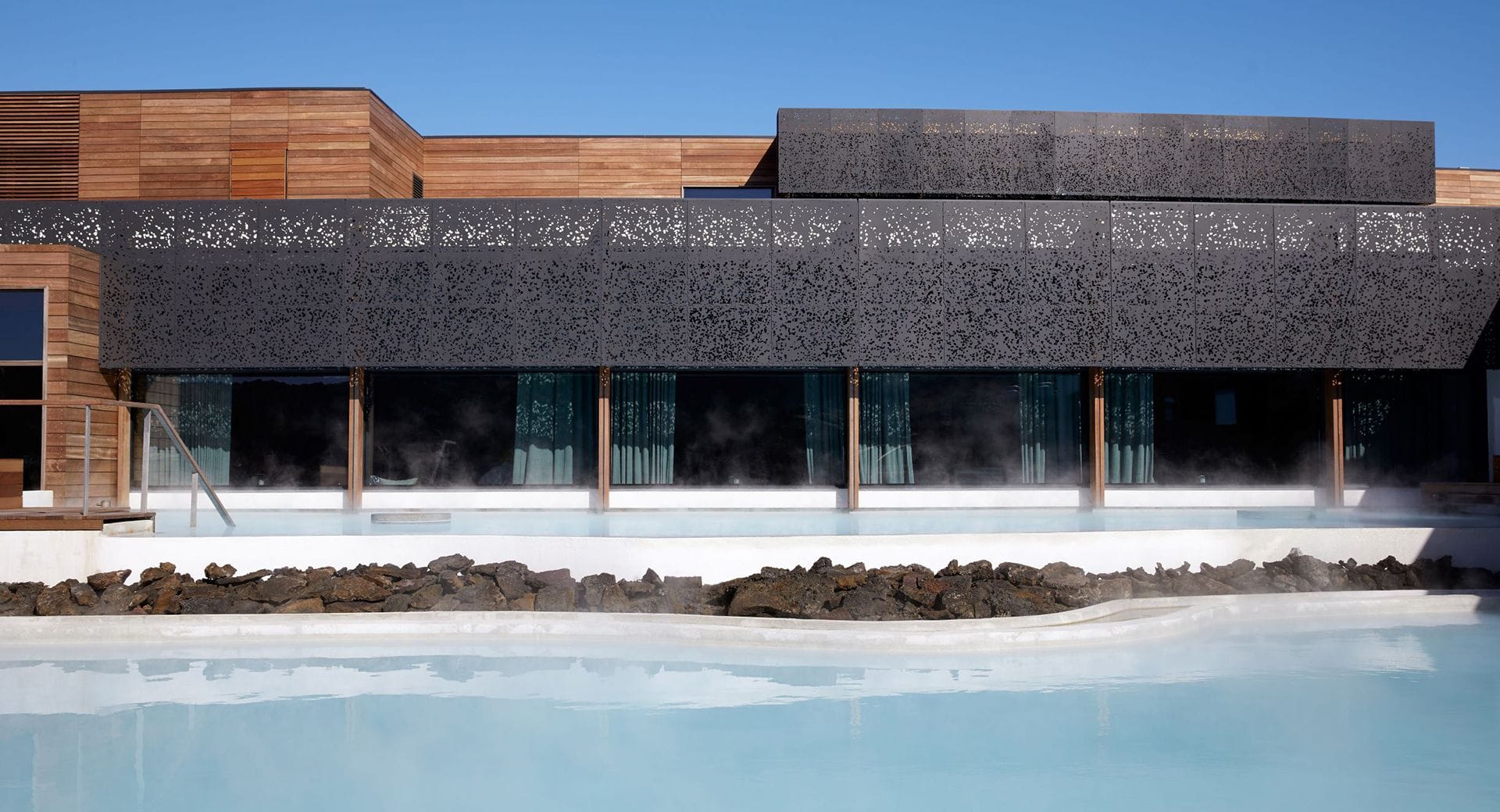 The front of the Blue Lagoon Dermatology Center building. The building is box-shaped and clad with both wood and black patterned panels. Windows are at the bottom of the building in a plane by the blue lagoon that lies right next to the building.