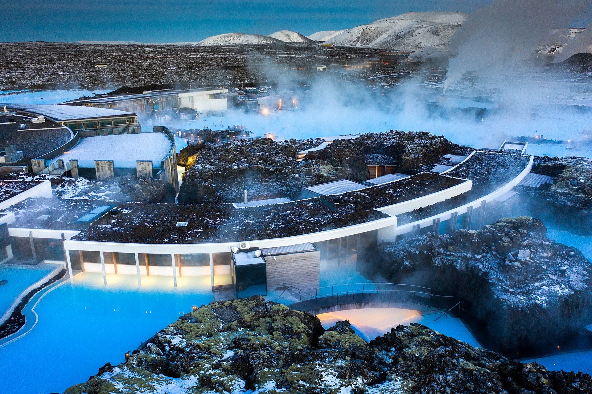 Arched structures surrounded by blue water and lava seen from above, with mountains in the background