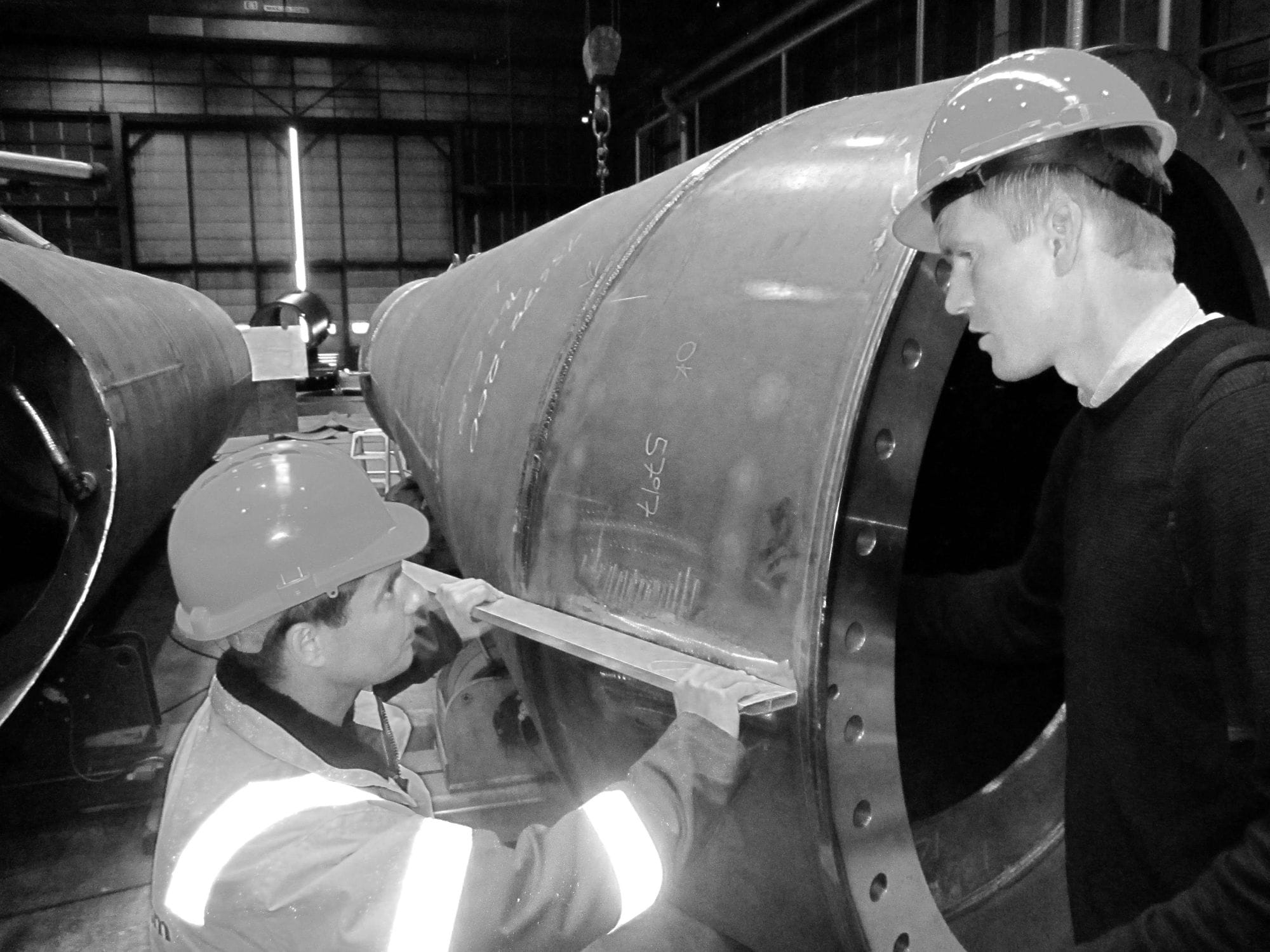 Grayscale photo af two men wearing security helmets looking at a mast in builiding