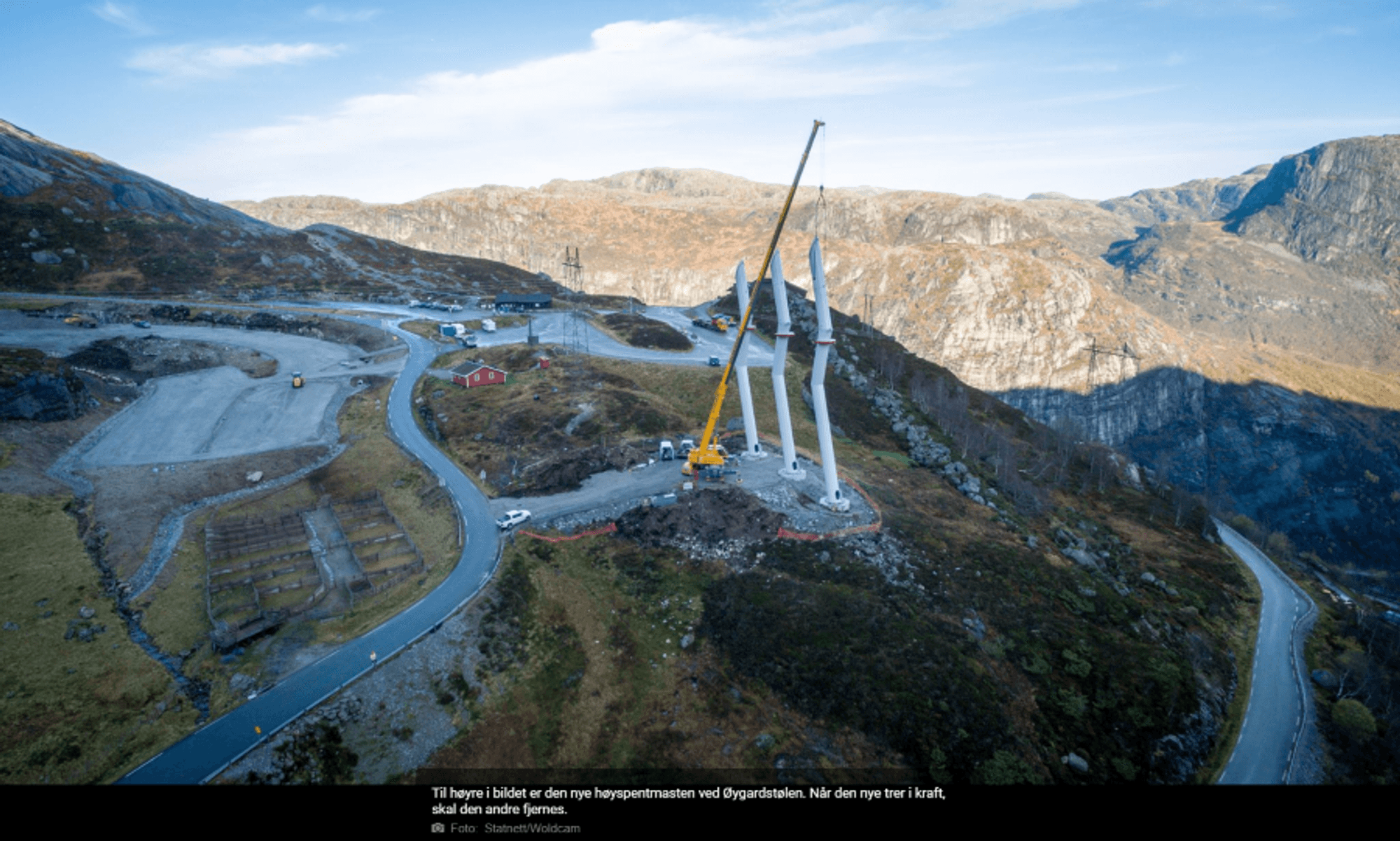 A buildingsite int he mountains with roads, cars and a crane putting up three white design masts