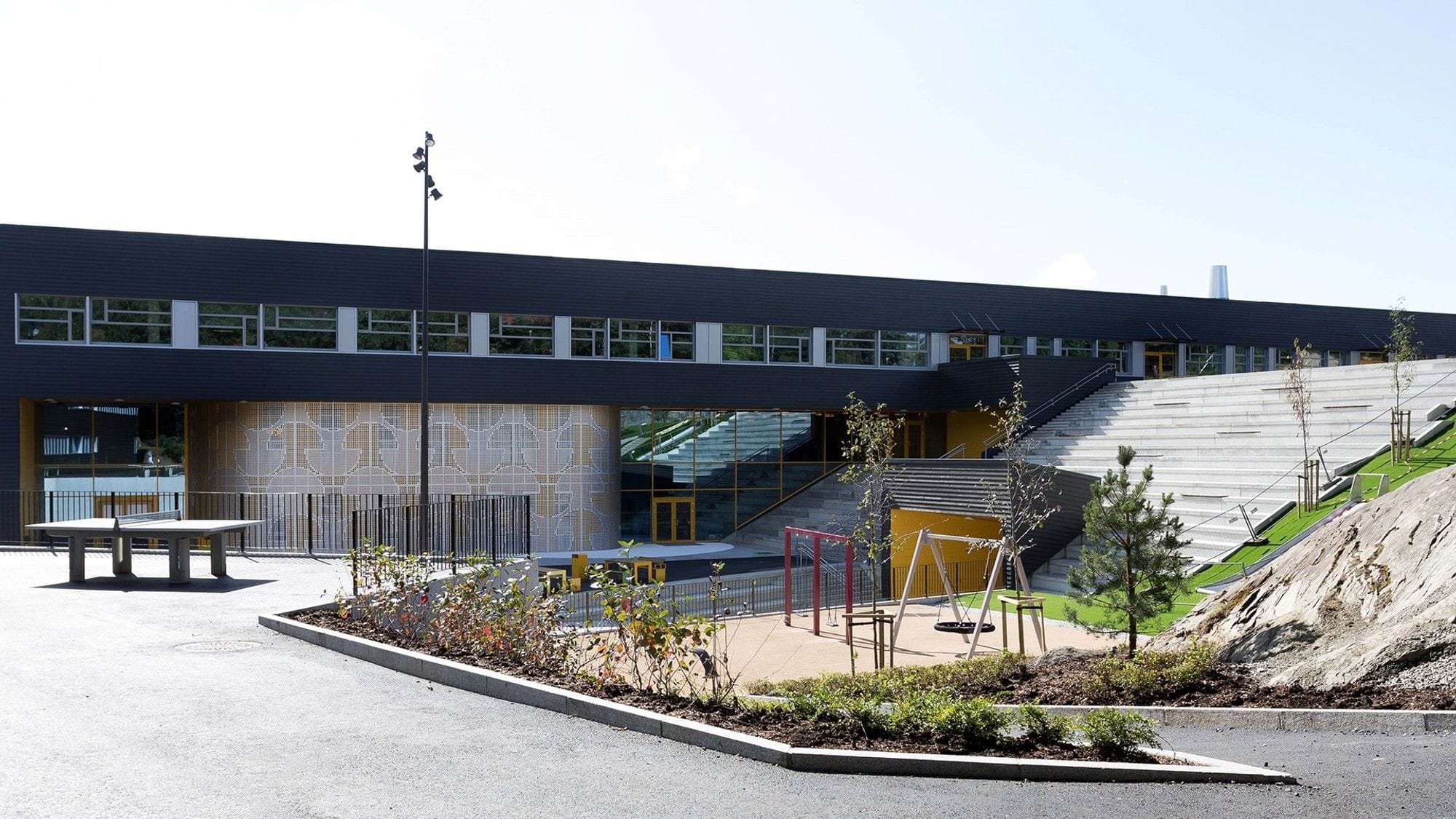 A plot with greenery, paved path, ping pong table and swings in front of a large building