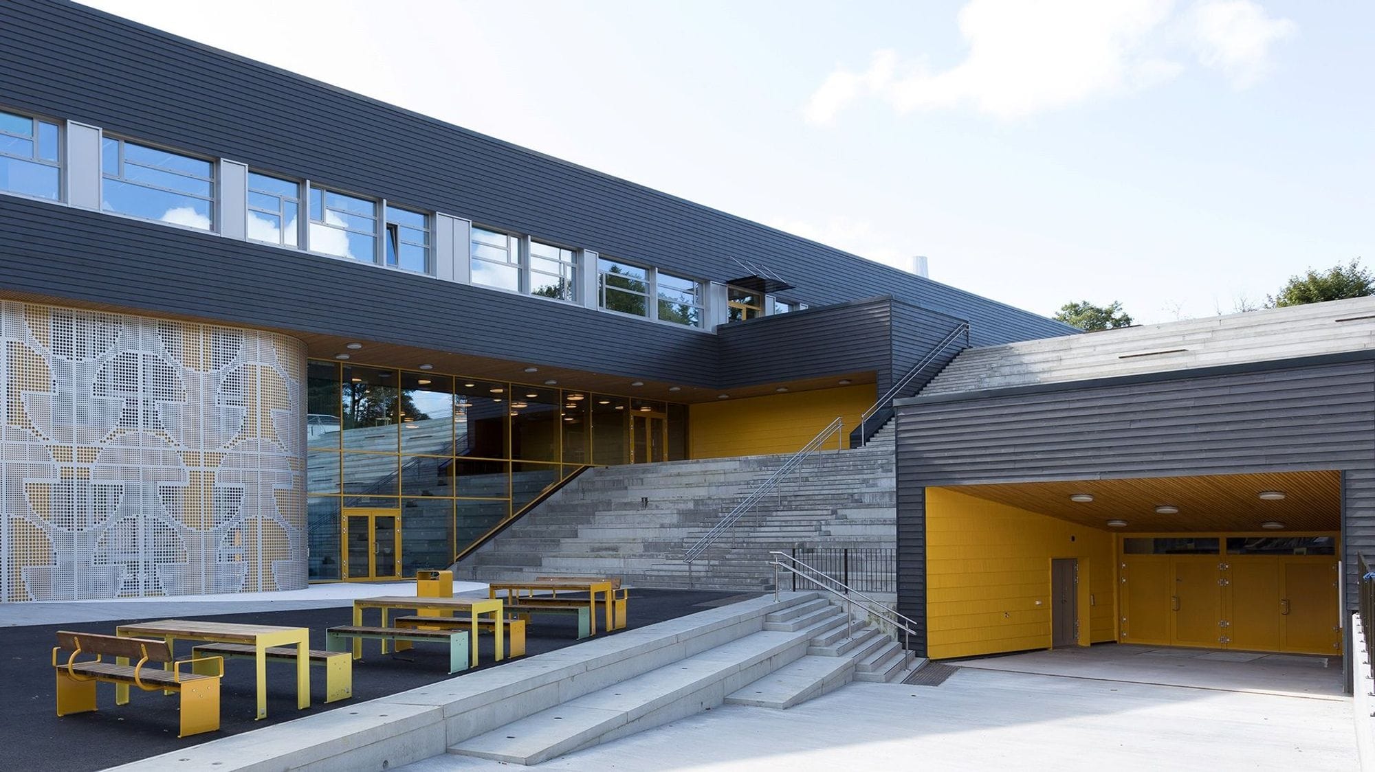 Concrete school lot in front of a black and yellow school building with benches in front of the building