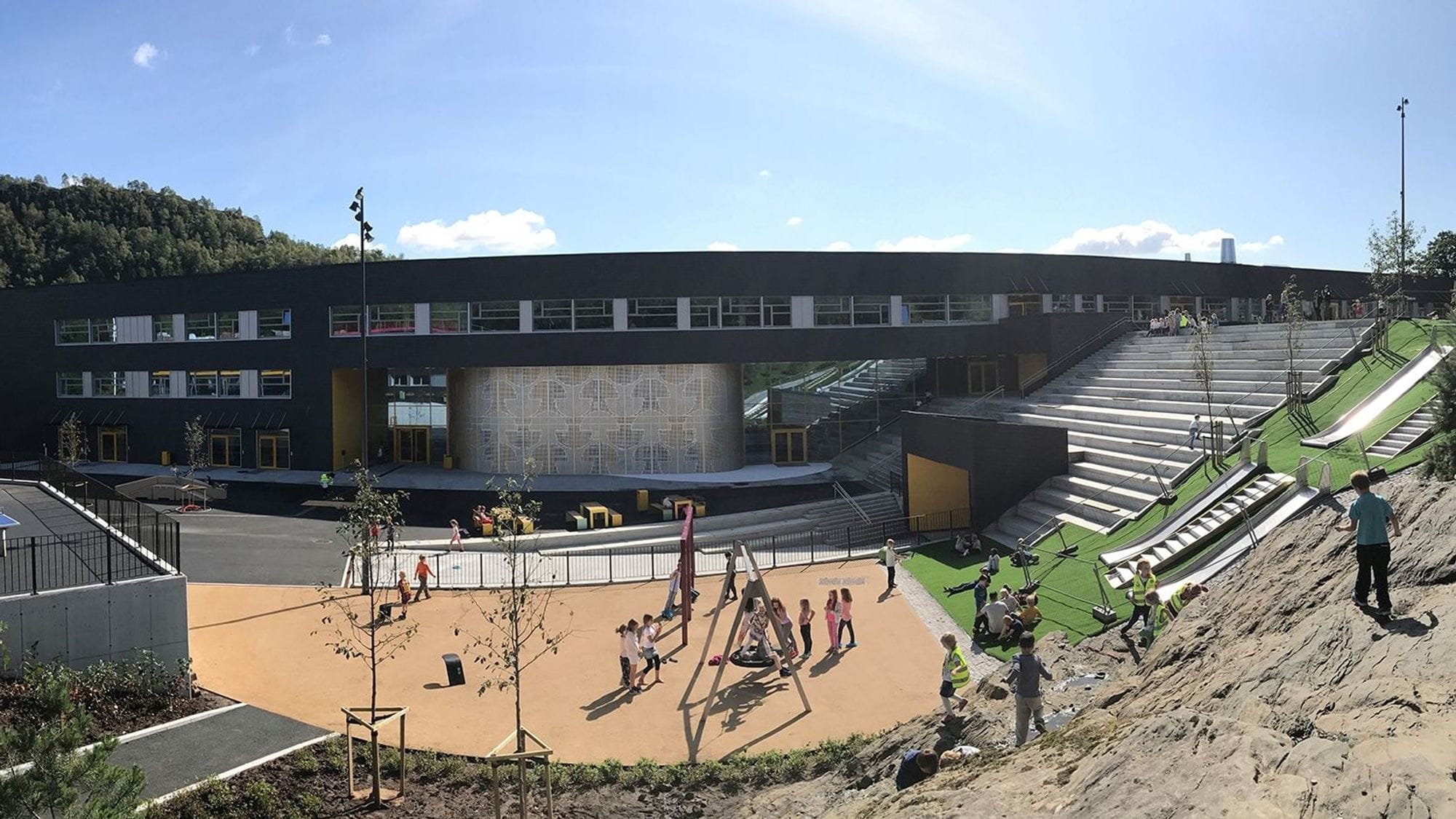 Overview of a school ode in front of a black school building. The plot has a playground with swings, slides on a green slope and a sloping wall with steps. Children are playing on the grounds.