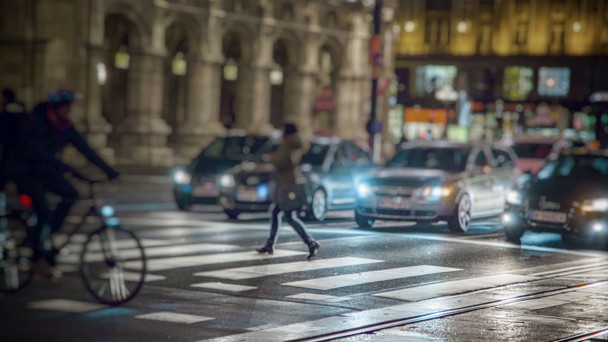 A traffic street in a big city, a crosswalk where a woman walks and a man rides a bicycle. It's dark outside and lights from cars and street lights create a mood