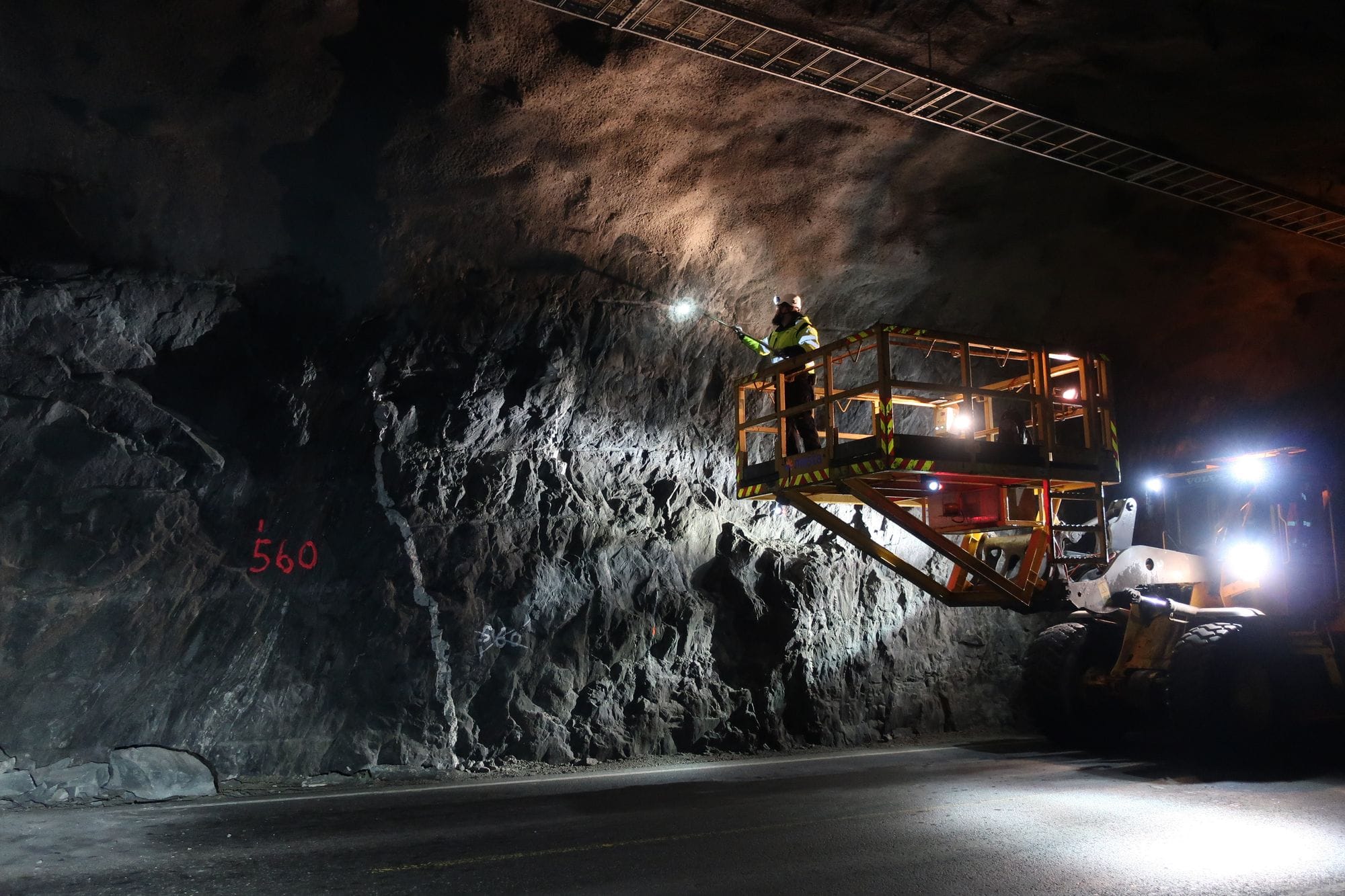 Photo taken in tunnels that have been dug into a rock or mountain. A man in safety gear stands on a crane and shines a light on the wall of the tunnel.